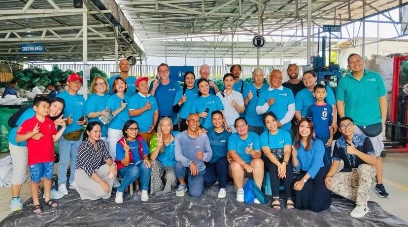 A large group of volunteers in matching blue shirts posing together at a Bali recycling facility.